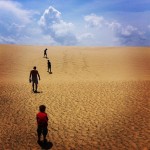 Four people wandering up a large sand dune with a blue sky and some clouds.